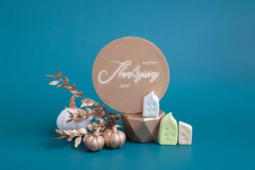 Still life of gold and white pumpkins, acorns and miniature houses on a turquoise background. Minimalistic autumn concept with text happy thanksgiving