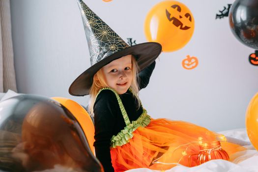 Children's Halloween - a girl in a witch hat and a carnival costume with airy orange and black balloons at home. Ready to celebrate Halloween.