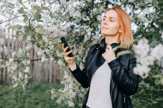 A red-haired teenage girl with headphones around her neck takes a selfie against the background of a flowering cherry tree. The time of spring flowering of cherry orchards.