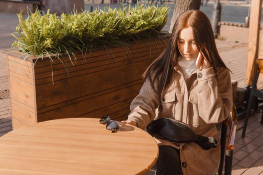 A stylish brunette woman in sunglasses is sitting at a table in an outdoor cafe with coffee and a phone. The girl is talking on the phone. Autumn walk.