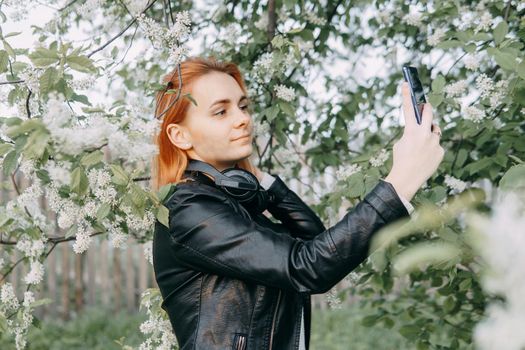 A red-haired teenage girl with headphones around her neck takes a selfie against the background of a flowering cherry tree. The time of spring flowering of cherry orchards.