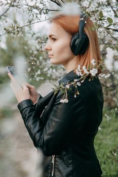 A red-haired teenage girl listens to music in headphones, configured via the phone. The time of spring flowering of cherry orchards.