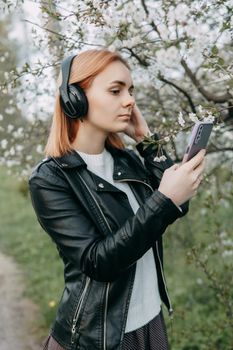 A red-haired teenage girl listens to music in headphones, configured via the phone. The time of spring flowering of cherry orchards.