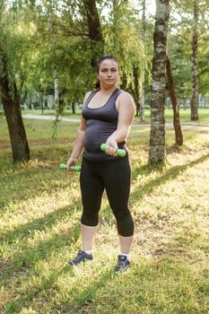 A charming brunette woman plus-size body positive practices sports in nature. Exercises with dumbbells.
