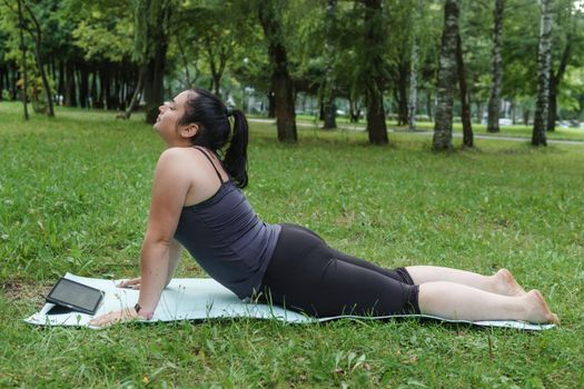 A charming brunette woman plus-size body positive practices sports in nature. Woman does yoga in the park on a sports mat. On a yoga mat in the dog pose.