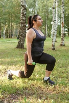 A charming brunette woman plus-size body positive practices sports in nature. Exercises with dumbbells.