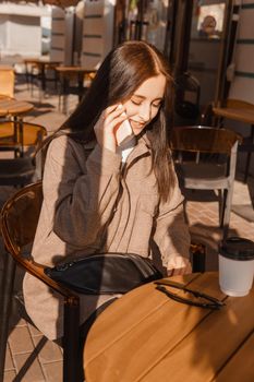 A stylish brunette woman is sitting at a table in an outdoor cafe with coffee and a phone. The girl is talking on the phone. Autumn walk.
