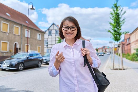 Portrait of smiling mature woman looking at camera on street of European small town. Positive middle aged lady in glasses shirt with bag walking outdoor. Age beauty vacation lifestyle people concept