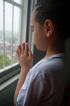 salvador, bahia, brazil - april 20, 2022: white ethnicity child praying next to an apartment window on a rainy day in the city of Salvador.