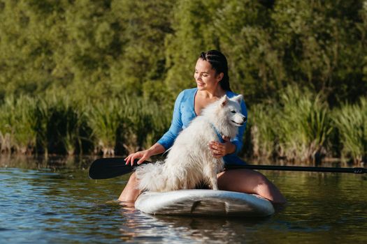 Happy Young Woman on the Lake at Early Morning Sitting on Sup Board with Her Dog Snow-White Japanese Spitz and Enjoying Sunrise