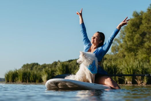Happy Young Woman Raising Hands Up While Sitting on Sup Board with Her Dog Japanese Spitz