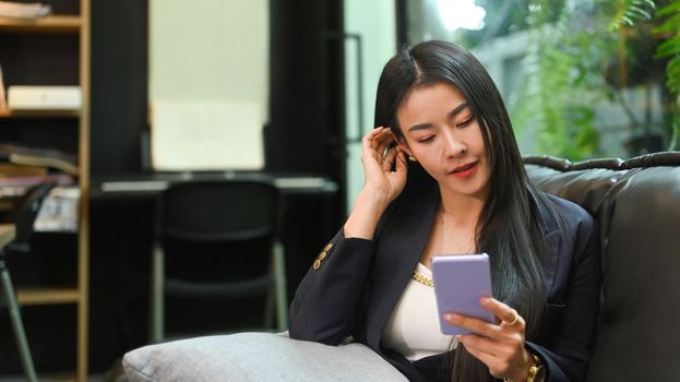 Asian working woman using mobile phone while resting on sofa at her personal office.