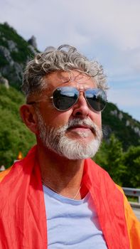 Portrait of a gray-haired senior elderly Caucasian man bisexuality with a beard and sunglasses with a rainbow LGBT flag in mountains. Celebrates Pride Month, Rainbow Flag Day