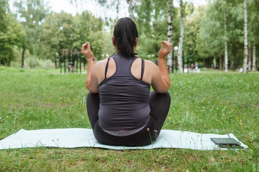 A charming brunette woman plus-size body positive practices sports in nature. Woman does yoga and meditation in the park on a sports mat