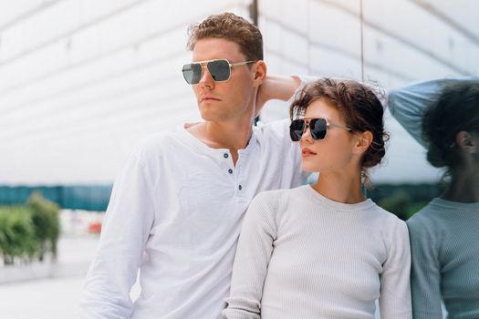 Smiling beautiful woman and her handsome boyfriend. Woman in casual summer jeans. Serious pensive couple in sunglasses walking business district. Couple posing on the street modern building background