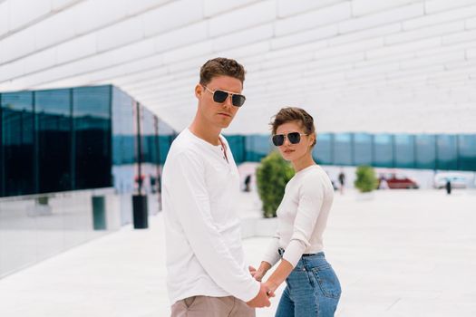 Smiling beautiful woman and her handsome boyfriend. Woman in casual summer jeans. Happy cheerful couple in sunglasses holding hand each other. Couple posing on the street modern building background