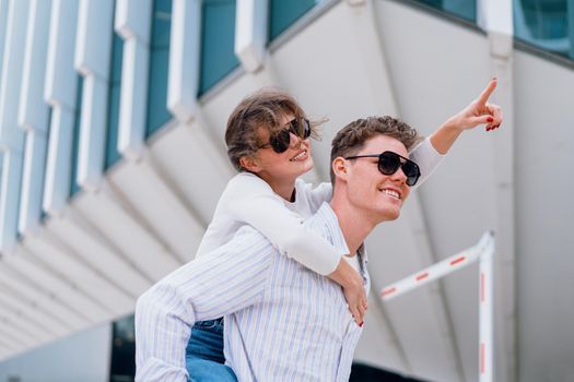 Carefree young urban couple in sunglasses doing piggyback at city street outdoor with modern commercial building on background. Man carrying young woman on his back, woman show by finger away