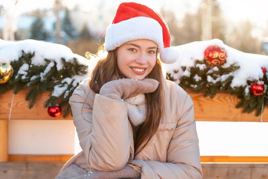 Winter portrait young adult beautiful woman in Santa hat. Christmas mood. Snowing. Winter beauty fashion concept. Pretty young woman sitting bench outdoor near Christmas garland