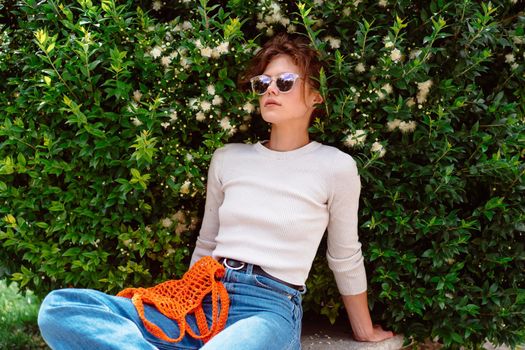 Young woman in light summer clothes with a reusable mesh eco bag and sunglasses, sitting bench at summer park before shopping. Sustainable lifestyle. Eco friendly concept.