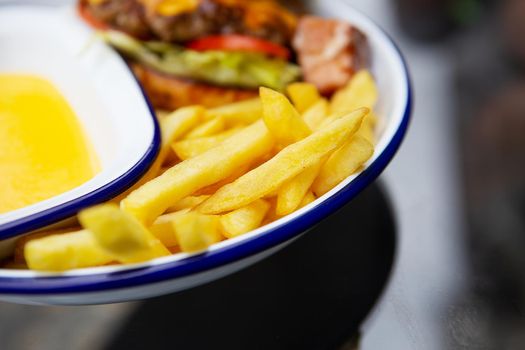 The concept of fast food. French fries lie on a meta plate along with cheddar cheese sauce and a hamburger in the background