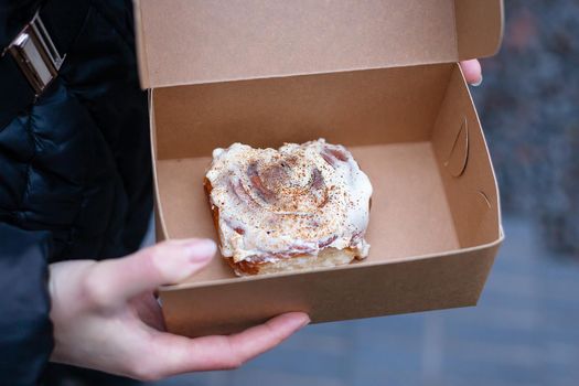 A girl holds a cinnabon with cream in a craft box. Fresh baked goods delivery