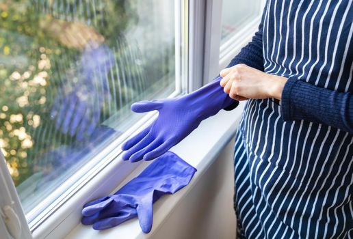 A young girl in a striped apron puts purple gloves on her hands. To prepare for washing windows in an apartment or house. Cleaning and cleaning concept