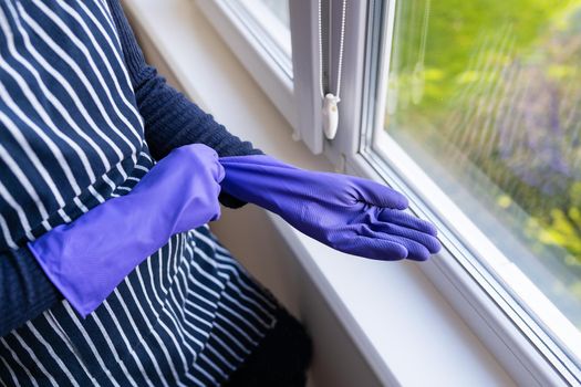 A young girl in a striped apron puts purple gloves on her hands. To prepare for washing windows in an apartment or house. Cleaning and cleaning concept