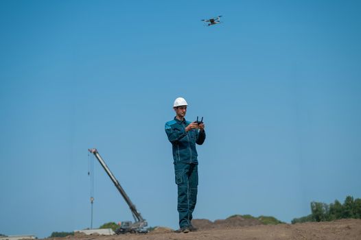 A man in a helmet and overalls controls a drone at a construction site. The builder carries out technical oversight