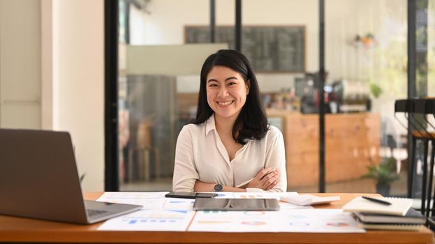 Confident asian woman employee sitting with arms crossed at her workplace and smiling to camera.