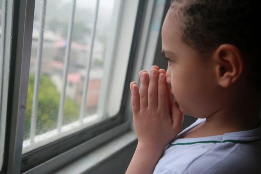 salvador, bahia, brazil - april 20, 2022: white ethnicity child praying next to an apartment window on a rainy day in the city of Salvador.