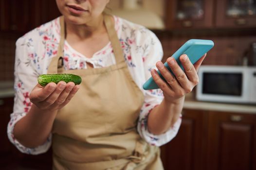 Cropped view of a housewife wearing a beige kitchen apron, looking through recipe of marinating cucumbers, in internet sites