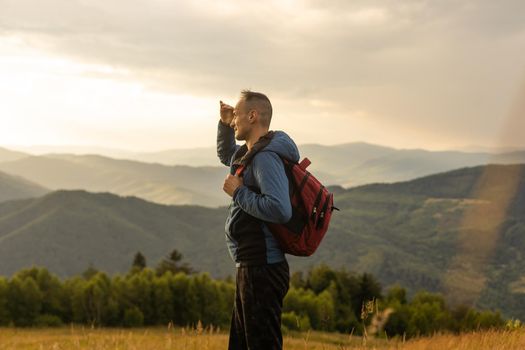 Guy tourist with a backpack stands in the mountains on a meadow with a backpack on his back.