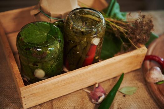 Still life. Jars of marinated chili peppers and pickled cucumbers, upside down on wooden board, with fresh ingredients and fragrant culinary herbs. Preparing homemade canned seasonal vegetables