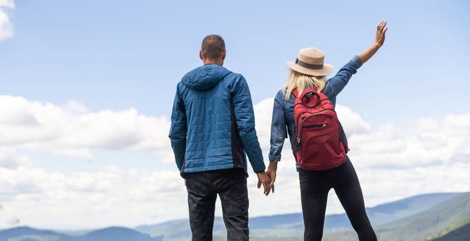 Rear view of hiking couple with backpack standing together on hill top enjoying beautiful landscape. Man and woman outdoors on hiking standing on a rock.
