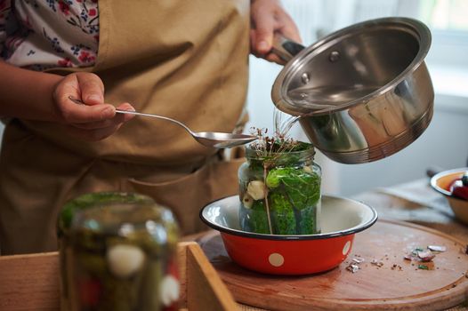 Close-up of a woman housewife in a chef's apron pouring boiling marinade over cucumbers in jars. Pickling cucumbers at home kitchen. Vegetable harvest conservation. Homemade canned food for winter