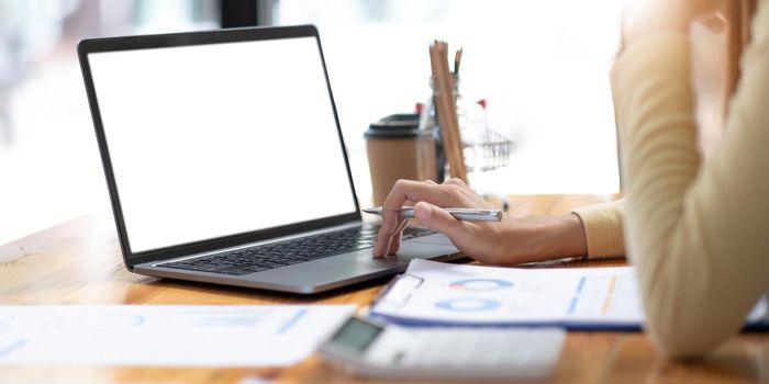 Mockup image of business woman using and typing on laptop with blank white screen and coffee cup on glass table in modern loft cafe.