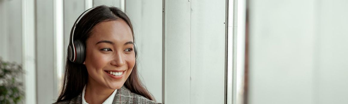 Asian Business woman listening music from phone in headphones standing near window in office