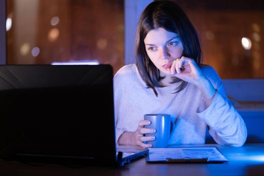 Young girl works at a laptop online at night against the background with the city lights outside the window. Woman is drinking coffee, holding a cup in her hands and reading the text on her device.