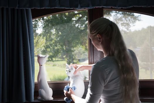 a woman wipes a porcelain figurine of a white cat on the windowsill,room cleaning and tidying up. High quality photo