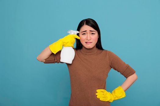 Frustrated tired young asian housemaid holding a spray near her temple isolated on a blue background, Cleaning home concept, Frightened sad female with negative state of mind