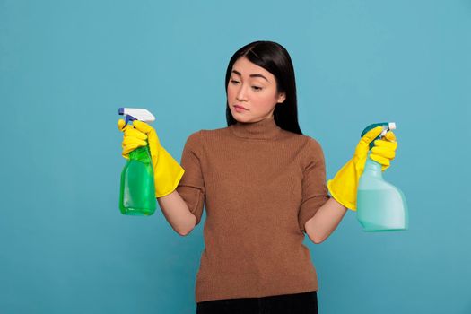 Portrait of pretty sad asian woman holding two detergent sprayers in yellow gloves isolated on a blue background, Cleaning home concept, Unhappy depressed and negative housewife
