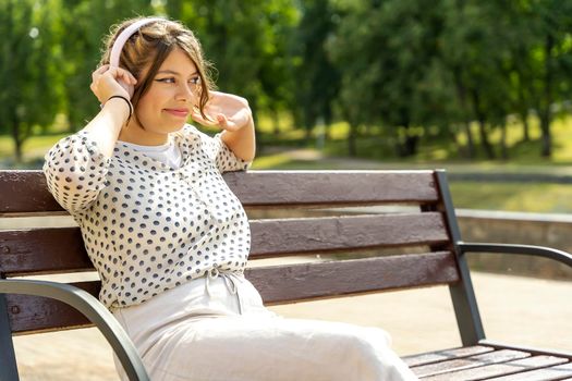 A cute Caucasian teenage girl in stylish clothes has a good time listening to music with headphones, sitting on a bench in a city park. a beautiful fashionable girl enjoys her free time