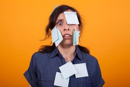 Portrait of cute young woman overwhelmed of work with sticky notes on her over yellow background in studio. Colorful sticky notes.