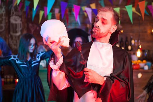 Attractive young man dressed up like dracula holding a black rose looking at his human skull at halloween celebration. Dracula costume.