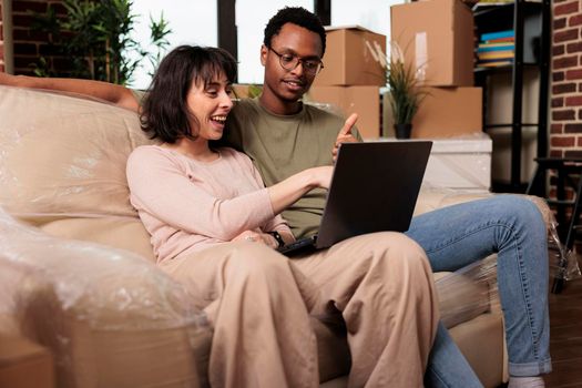 Diverse couple doing online shopping on laptop to buy furniture, browsing internet website for home decor inspiration. Moving in rented apartment property together for relationship event.