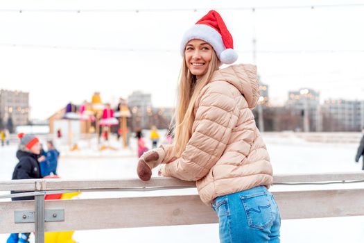 Outdoor portrait young beautiful fashionable woman wearing Santa hat, posing street of European city. Winter Christmas holidays concept. ice skate rink background. Lean on fence near ice skate rink