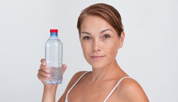 Portrait of caucasian smiling middle aged woman holding plastic bottle of water with screw top on white background
