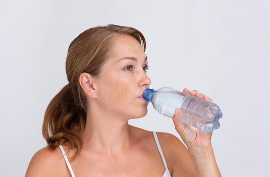 Beautiful caucasian middle aged woman drinking pure water from plastic bottle on white background looking aside