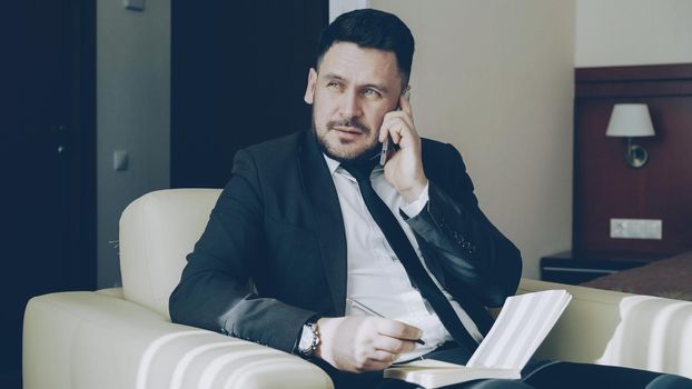 confident businessman talking mobile phone and writing notes in notepad while sitting on armchair in hotel room. Travel, business and people concept