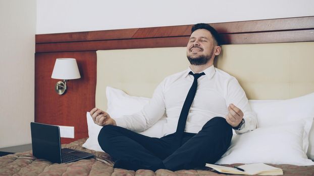 Happy businessman put off laptop computer and taking yoga lotus position sitting on bed in hotel room. Travel, business and people concept